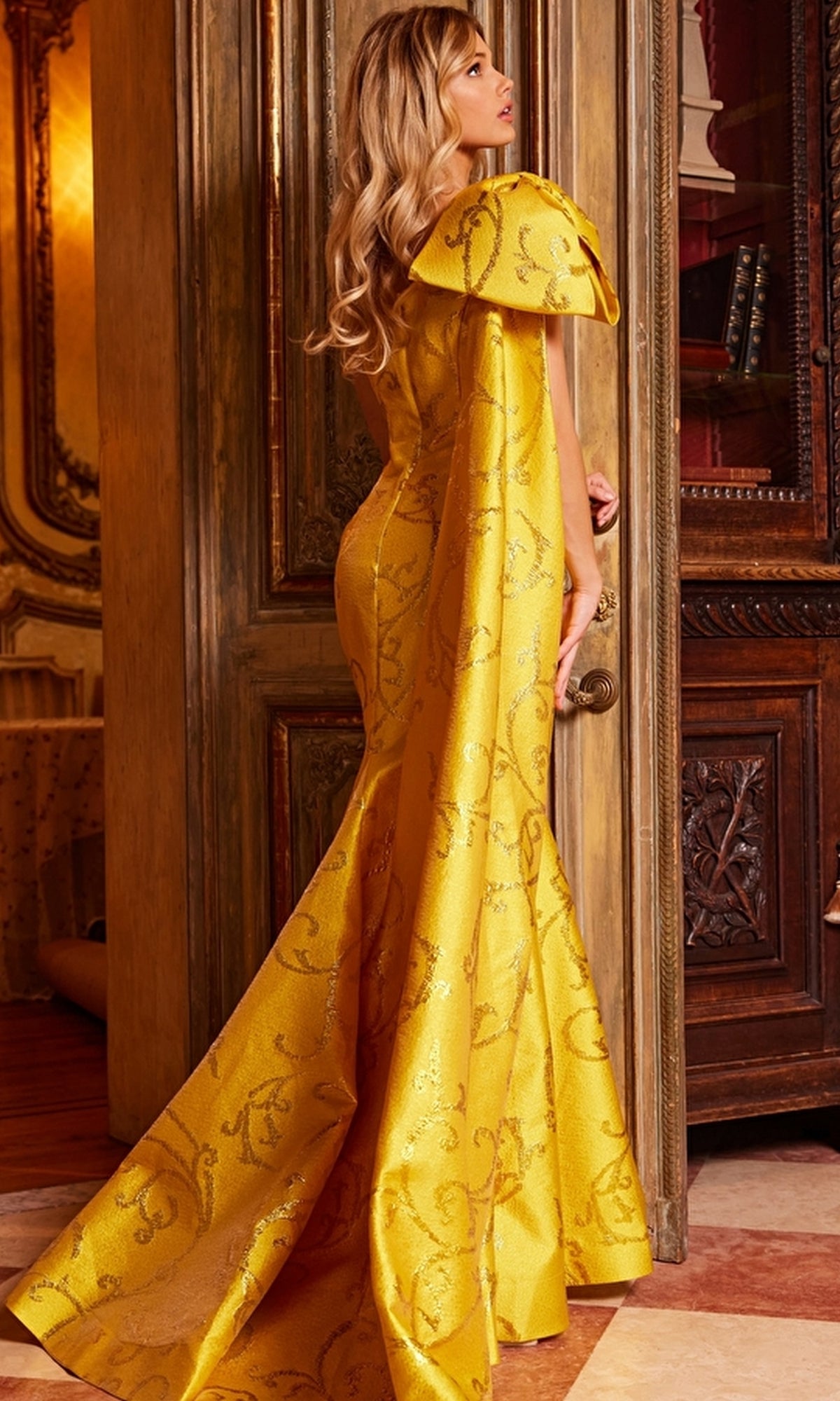 A woman in a fitted, floor-length yellow Jovani One-Shoulder-Bow Long Formal Dress 23742 with a train stands before an ornate wooden door.
